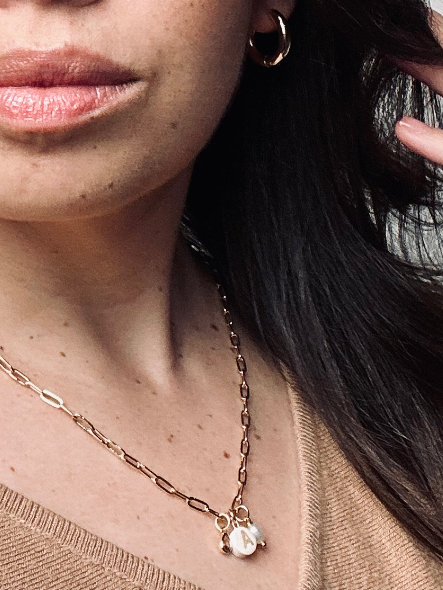 Personalized clay initial necklace from the “Your Story with Grace” collection worn by a model, paired with gold hoop earrings. Features a white round polymer clay charm with a gold initial, accented with a clear teardrop crystal and a fresh water pearl, hanging on an 18k gold-filled paperclip chain. The look radiates elegance, individuality, and timeless self-expression.