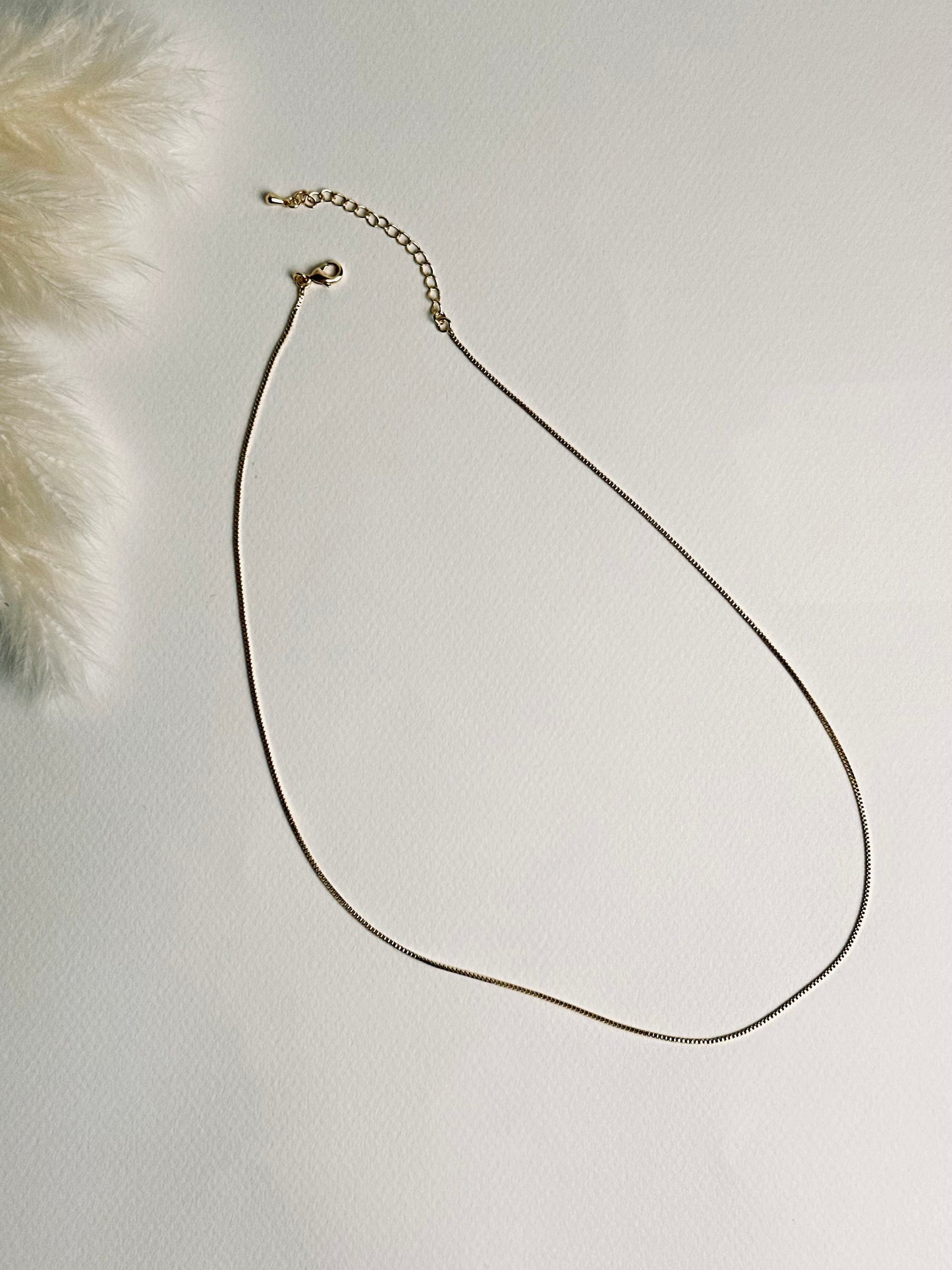 Serena — Gold Box Chain Necklace from the Timeless Grace collection, a thin gold-plated brass chain displayed on a book with focus on the lobster clasp and 2-inch extender. The delicate links and adjustable design highlight both elegance and practicality, symbolizing versatility, grace, and effortless style.