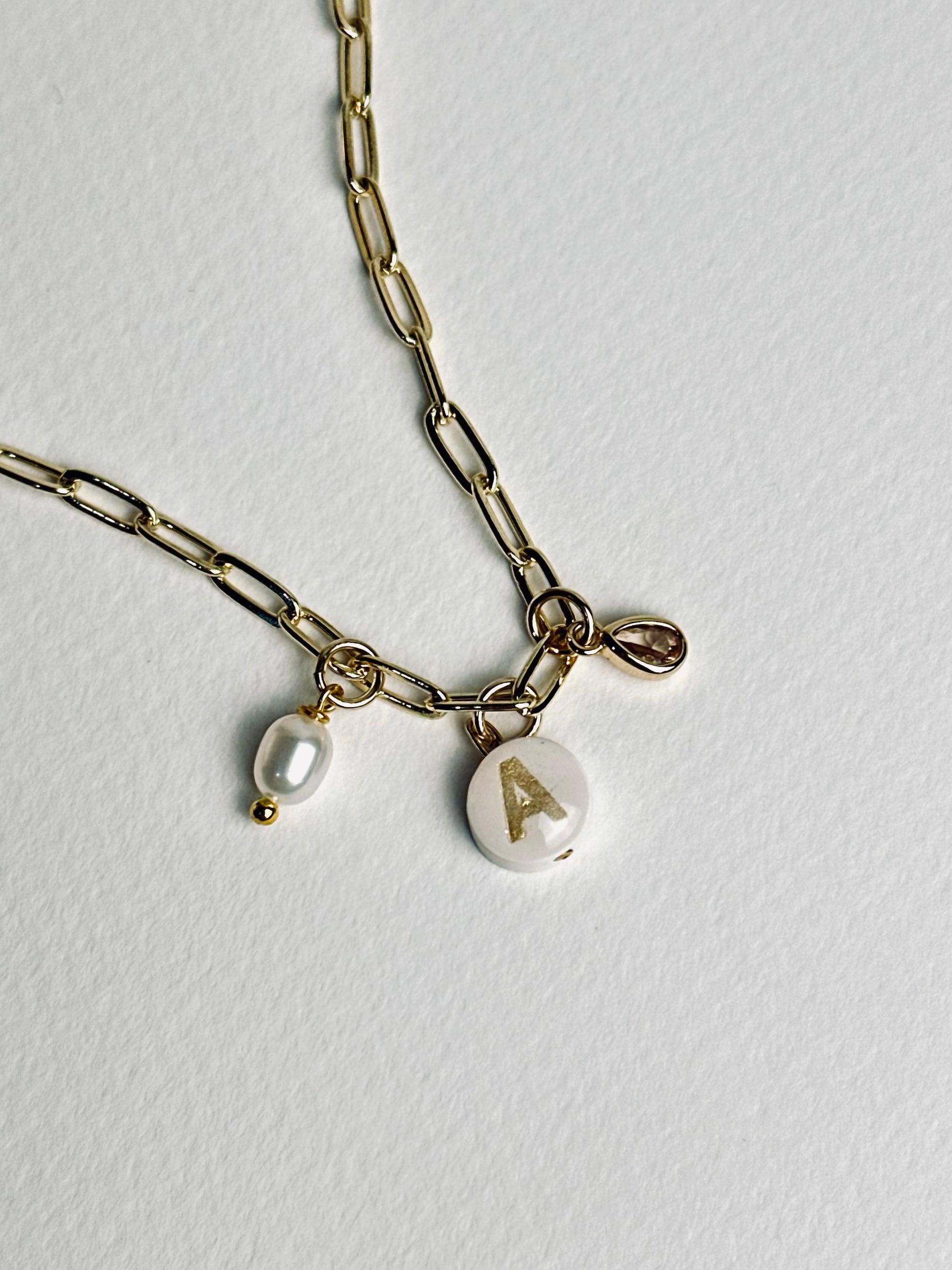 Close-up of the charms on the personalized clay initial necklace from the “Your Story with Grace” collection, showing the white round polymer clay charm with a gold initial, the clear teardrop crystal, and the freshwater pearl. The detail highlights elegance, individuality, and delicate craftsmanship.
