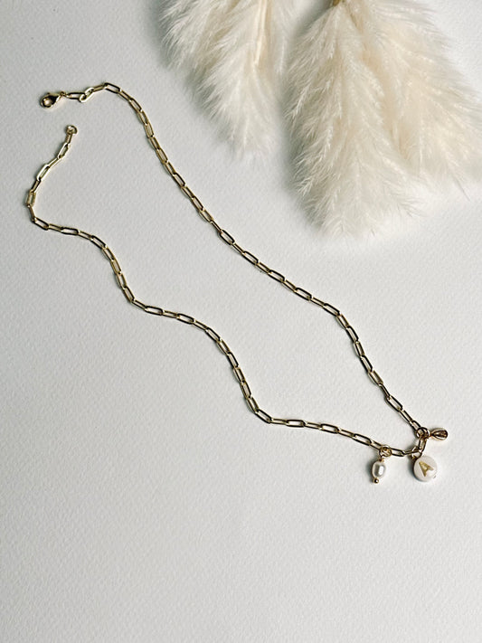 Personalized clay initial necklace from the “Your Story with Grace” collection laid flat, featuring a white round polymer clay charm with a gold initial, accented with a clear teardrop crystal and a freshwater pearl, on an 18k gold-filled paperclip chain. The design highlights elegance, individuality, and meaningful self-expression.