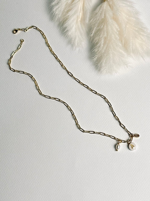 Personalized clay initial necklace from the “Your Story with Grace” collection laid flat, featuring a white round polymer clay charm with a gold initial, accented with a clear teardrop crystal and a freshwater pearl, on an 18k gold-filled paperclip chain. The design highlights elegance, individuality, and meaningful self-expression.