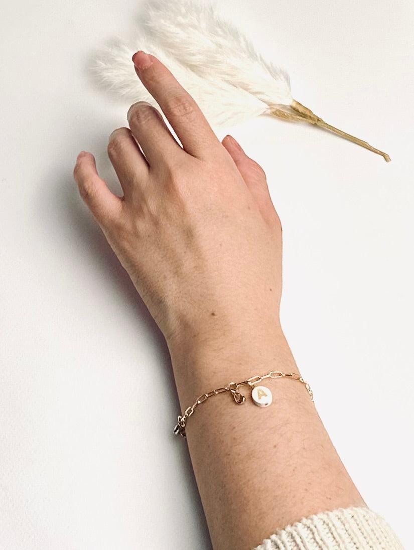 Personalized clay initial bracelet from the “Your Story with Grace” collection worn on a model’s wrist. The white polymer clay charm with a gold initial hangs from a 24k gold-filled paperclip chain with a sparkling crystal teardrop, highlighting elegance, individuality, and meaningful self-expression.