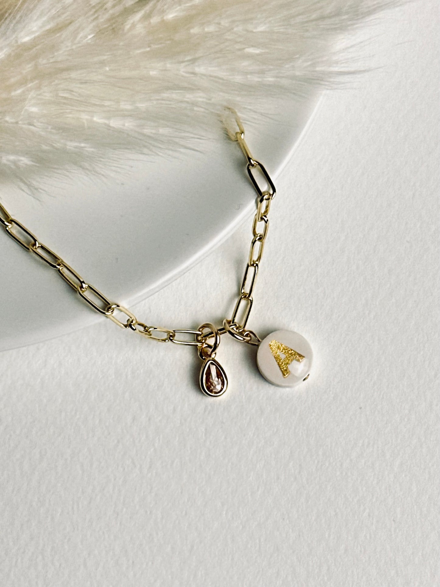 Personalized clay initial bracelet from the “Your Story with Grace” collection, laid flat. Features a white round polymer clay base with a gold initial, suspended on a 24k gold-filled paperclip chain and accented with a sparkling crystal teardrop, embodying elegance, individuality, and meaningful self-expression.