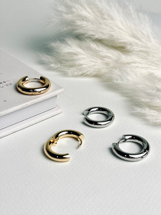 Radiance Hoop earrings from the “Timeless Grace” collection, 20mm thick hoops displayed flat with clasps open, showing both gold and silver options. Their bold, polished design reflects strength and elegance, inviting a sense of confidence and timeless beauty.