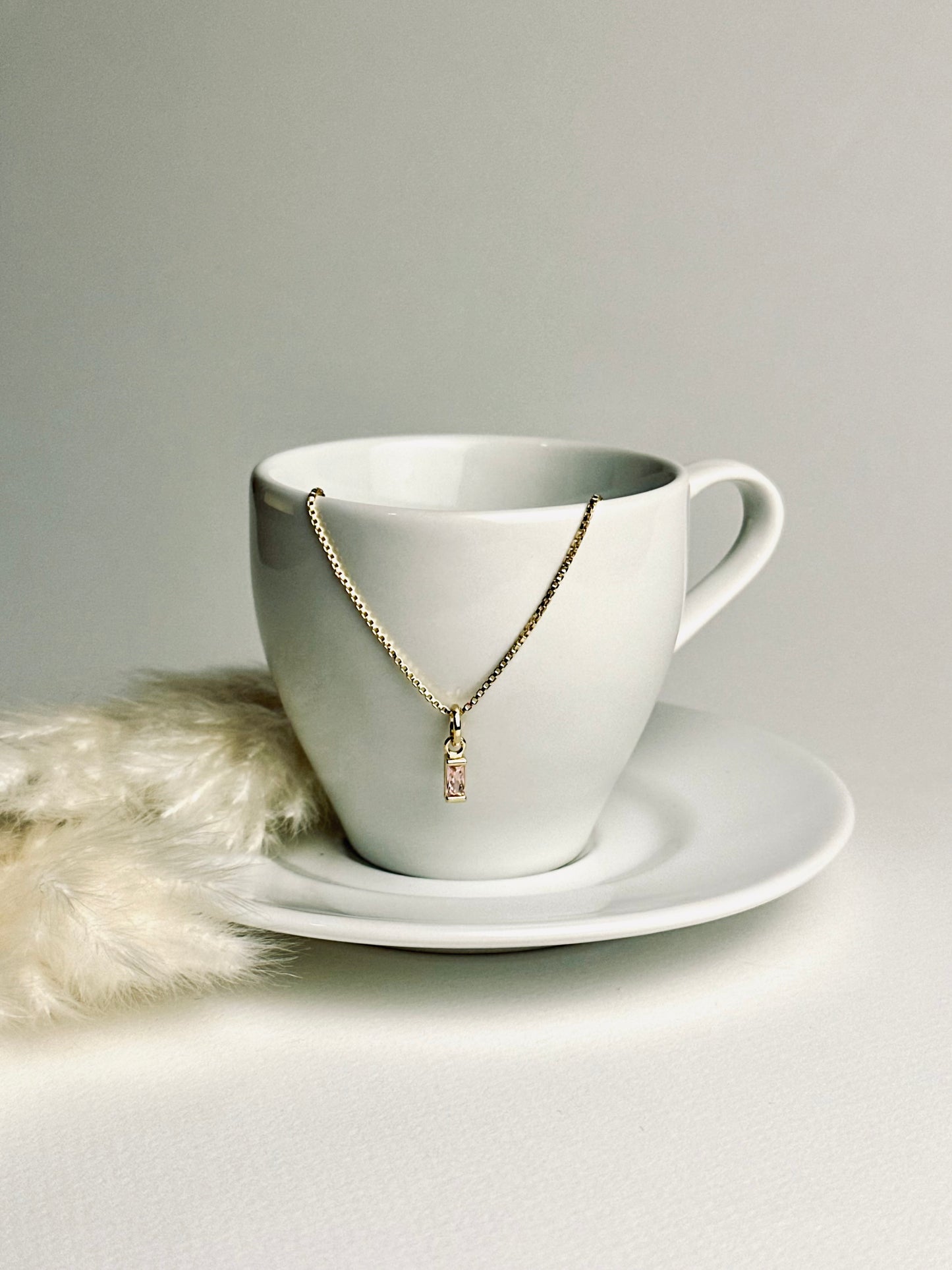Aesthetic photo of a birthstone necklace delicately dangling over an espresso cup, with soft feather pampas in the background. The warm tones and gentle light evoke calm, elegance, and the meaningful beauty of everyday moments.